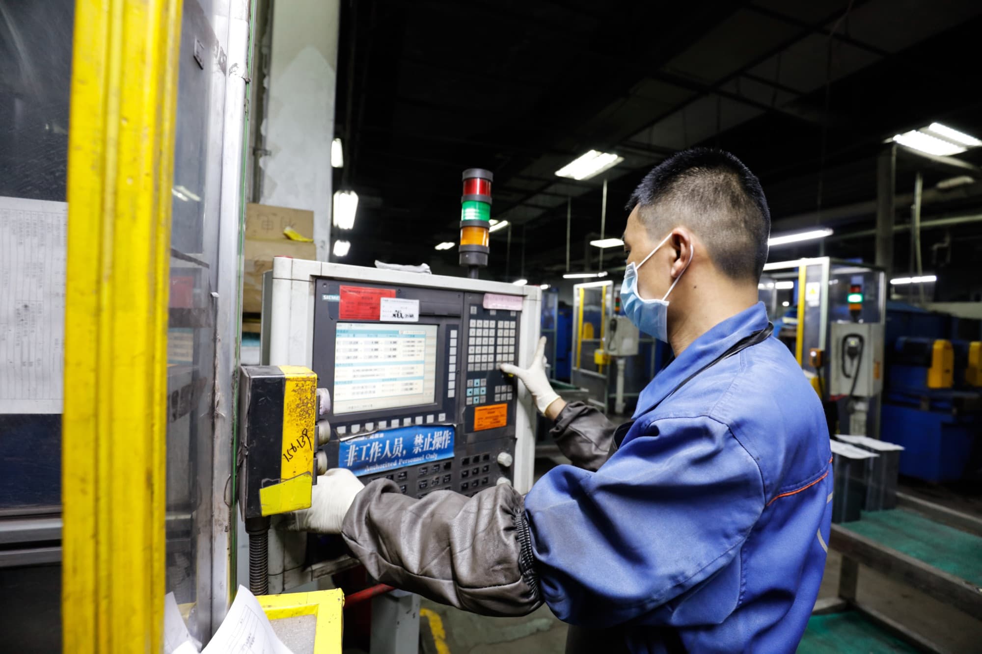China supplier factory — masked operator working at a CNC machine control panel during a Sinospect factory visit; production line and signal tower visible in the background.