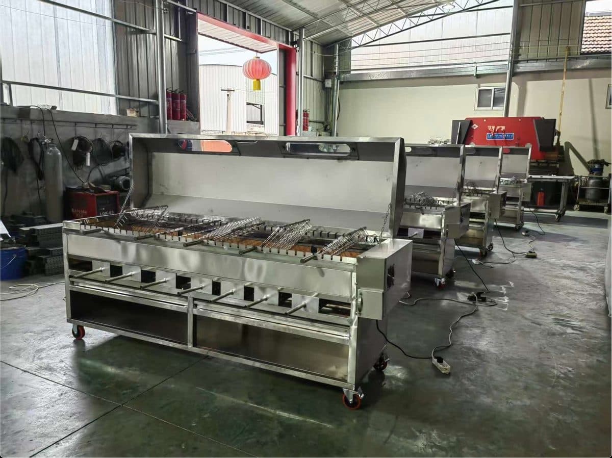 Stainless steel charcoal rotisserie pits with hoods and flat baskets on casters, photographed at the workshop