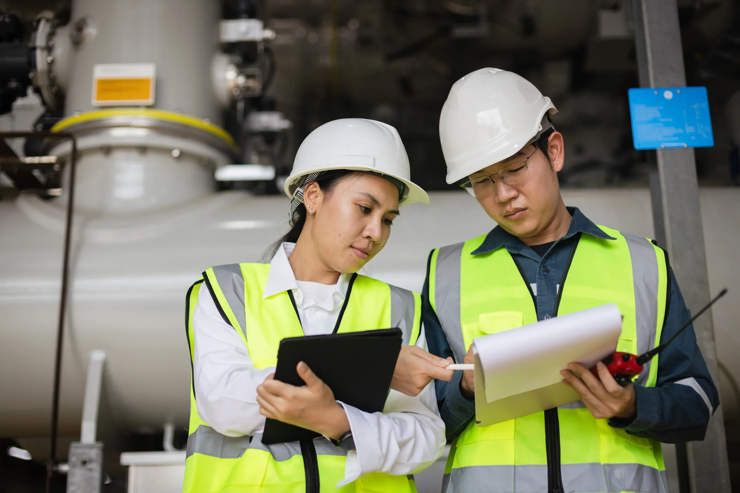 Two engineers in hi-vis vests and hard hats cross-checking documentation against equipment during a factory acceptance test